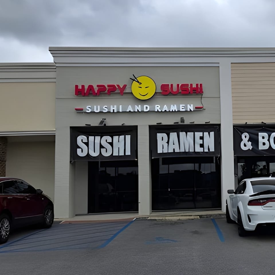 Exterior of Happy Sushi restaurant with red lettering and a yellow smiley face logo, two cars parked outside.